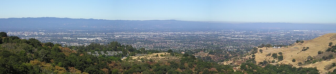 A panorama looking west across the Santa Clara Valley from the hills of Alum Rock Park: oak woodland and dry grass hills in the foreground, the cities spread across the valley floor, and the Santa Cruz Mountains in the haze beyond